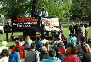 Photo of demonstration. Crowd seated on grass facing speakers on platform with large banners: 'Haringey Black Action: Black Lesbians and Gays Fight Back!' and 'Fight Bigotry, Smash the Backlash, Fight Racism'.
