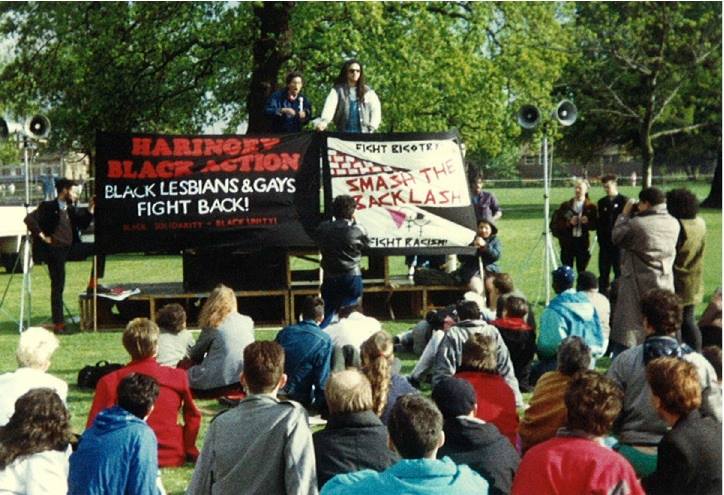 Photo of demonstration. Crowd seated on grass facing speakers on platform with large banners: 'Haringey Black Action: Black Lesbians and Gays Fight Back!' and 'Fight Bigotry, Smash the Backlash, Fight Racism'.