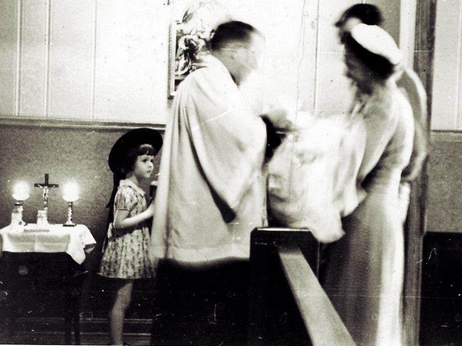 Black and white photo, looks to be 1950s or 1960s. In a church, during a baptism. A priest stands facing a mother and father, the mother holds the baby. There is a young girl looking on. The image is blurred where the priest and baby are moving.