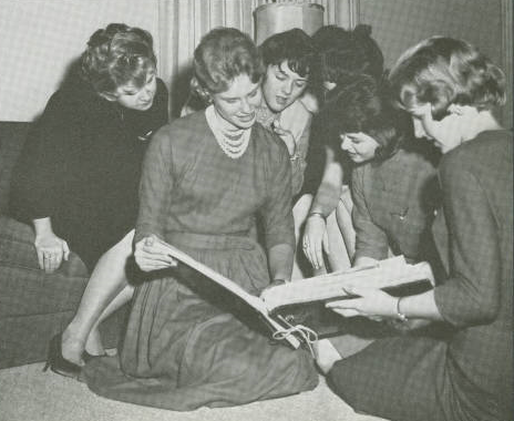 Black and white photo of group of young women looking through a book. They are seated or kneeling on the floor. Smiling and interested. 
