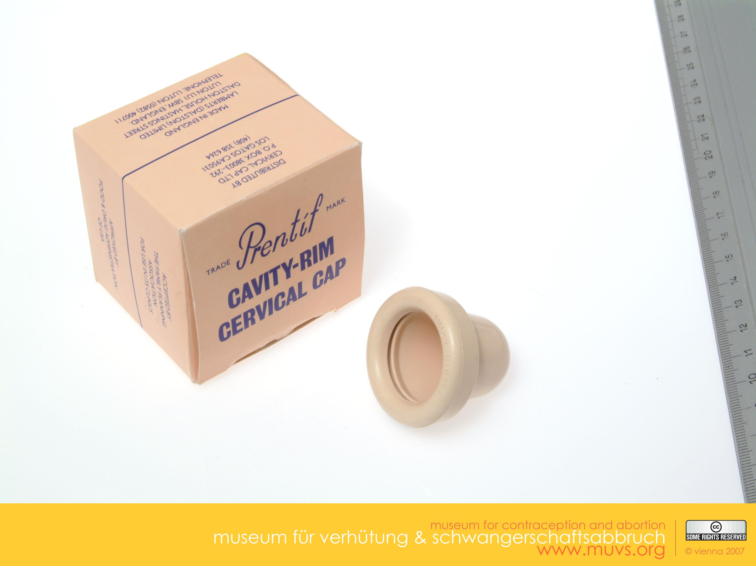 Photograph of the Prentif Cavity Rim Cervical Cap, with packaging. Ruler alongside shows rough size. The cap is beige, teat shaped, hollow, about 3 cm long, rim about the same diameter. Box is beige, square, with branding, blue writing. The Prentif Cavity Rim Cervical Cap was one of the cap types that the FDA approved in 1988 for American women’s use. Courtesy of the Museum of Contraception and Abortion, Vienna, Austria (MUVS, Wien).