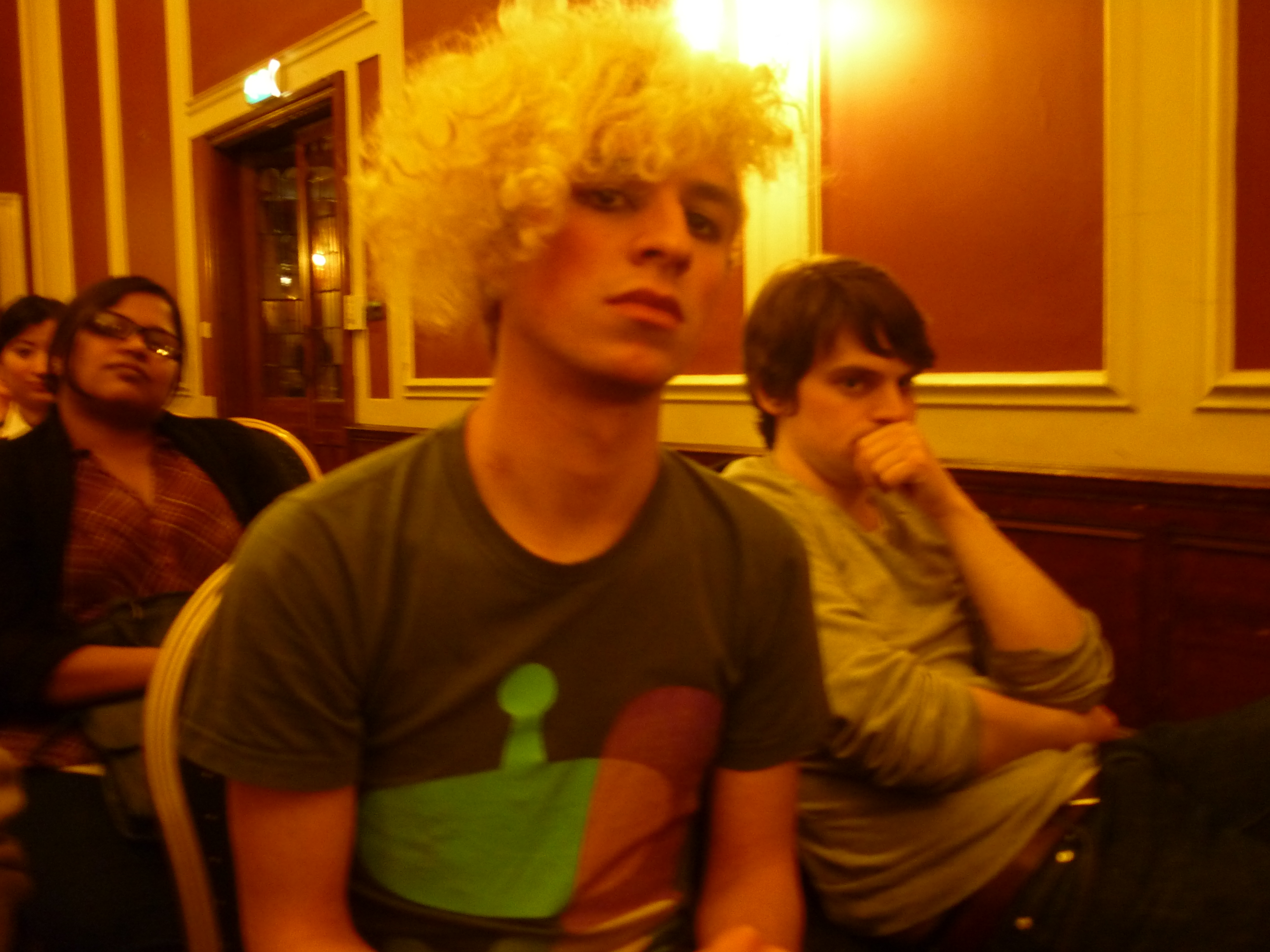 Photo taken at event. Young person, 20s, looks into camera with attitude and a look of defiance. Wearing T-shirt and with a mop of dyed blonde, curly hair. A second young person sits behind. Hand covering part of face. Looks sideways into camera