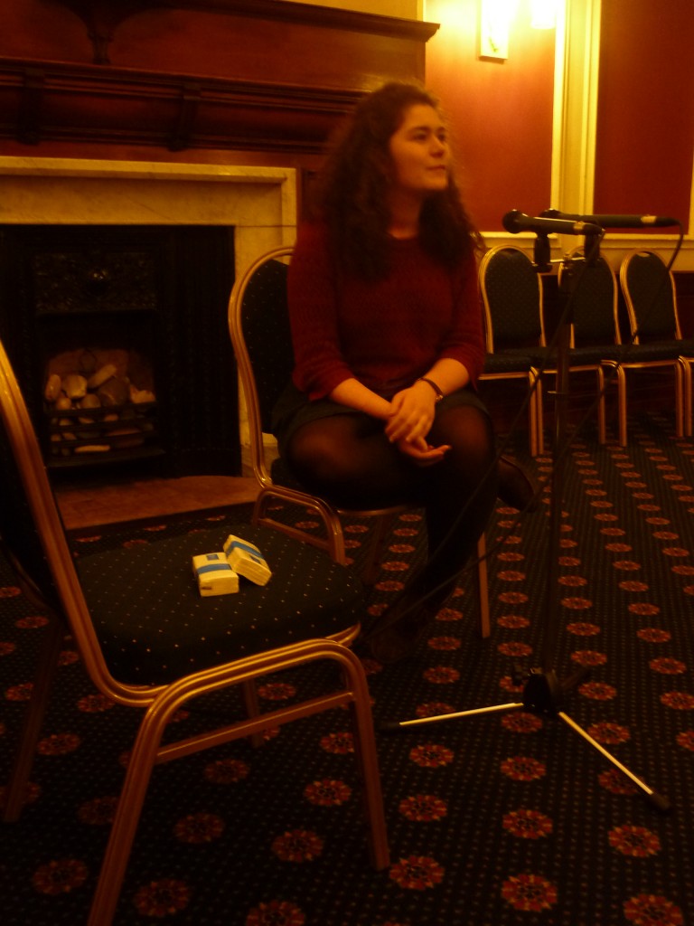Young woman sits talking into microphone in front of her. She is in a function room. Fireplace behind her. 