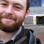 Head and shoulder view of Gareth Smith standing in front of building with EU flag over door. Gareth has a beard and is smiling. He is wearing a jacket and backpack