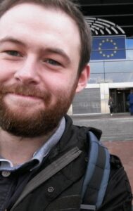 Head and shoulder view of Gareth Smith standing in front of building with EU flag over door. Gareth has a beard and is smiling. He is wearing a jacket and backpack