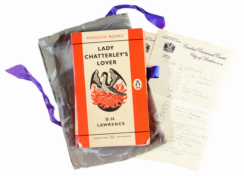 Birdseye photo of Penguin paperback edition of Lady Chatterley's Lover and handwritten trial notes on headed Central Criminal Court paper. The novel's cover has a line drawing of a phoenix rising from flames. Sitting on purple ribbon and grey packet.