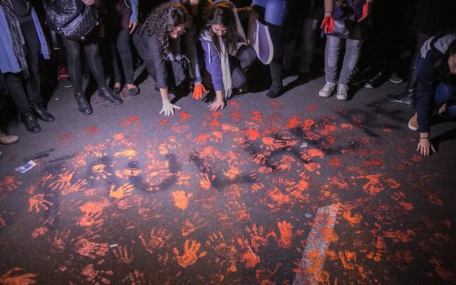 Orange handprints cover a section of pavement. About a dozen people are in view from the waist down, several of them with painted hands on the ground.
