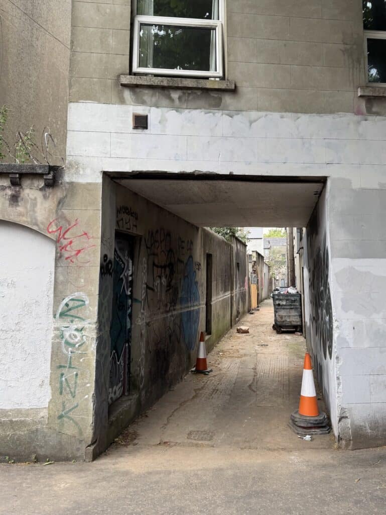 A secluded alleyway. Several orange cones and graffiti are visible.