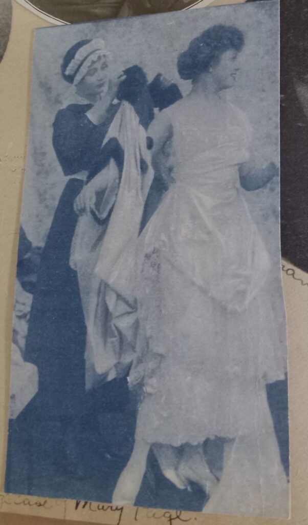 A woman in a beautiful white gown being dressed by another woman in dark plain clothes