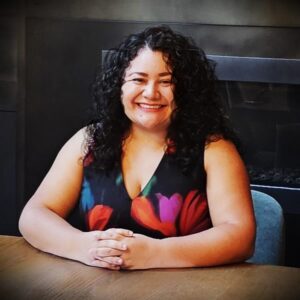 Isabel Millan seated at a table, smiling, with long back curly hair